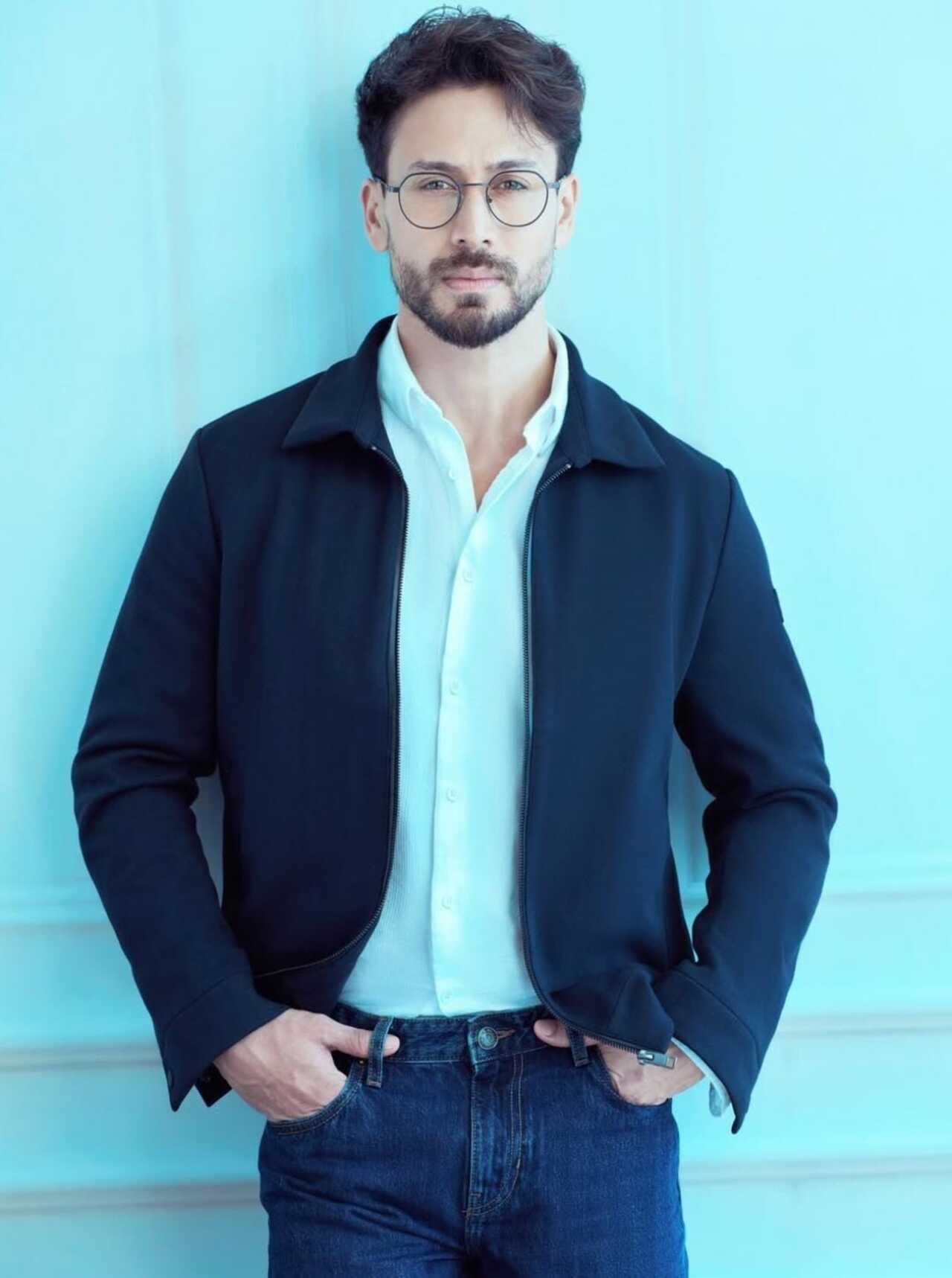 Tiger dons a polished navy zip-up jacket over a crisp white shirt and dark jeans, completed with round wire-rimmed glasses. This sophisticated attire steps away from his usual glam outfits.